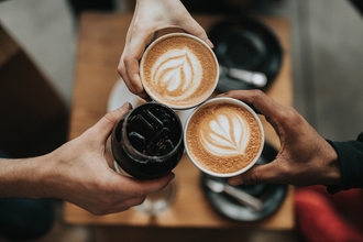 Camera looking down on three hands holding coffee cups together. Cups are filled with coffee, two with a leaf foam pattern.