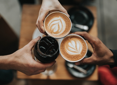 Camera looking down on three hands holding coffee cups together. Cups are filled with coffee, two with a leaf foam pattern.